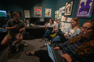 Four people sit in a radio control room. They are talking and using computers.