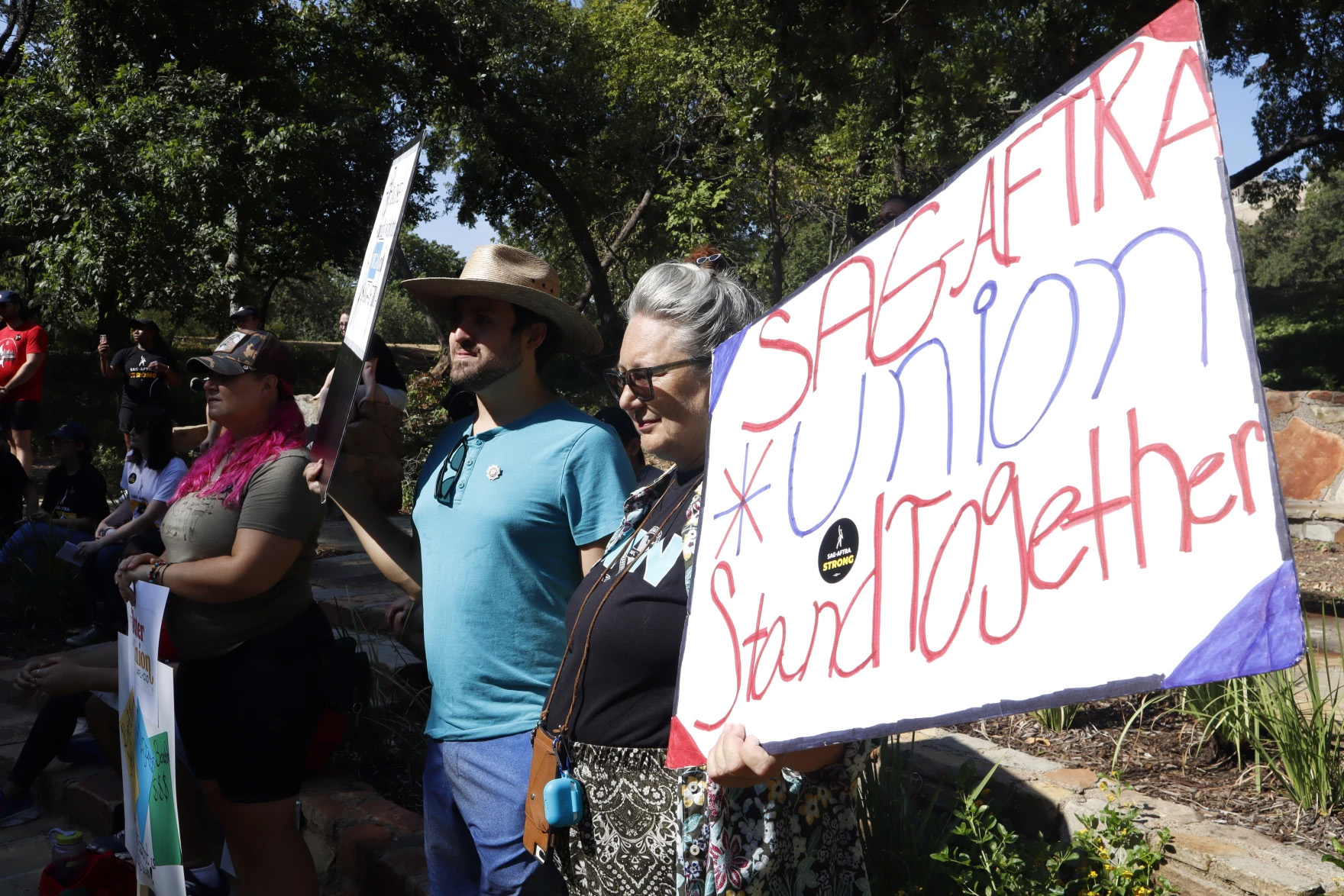 A group of SAG-AFTRA union members gather holding picket signs. One reads 