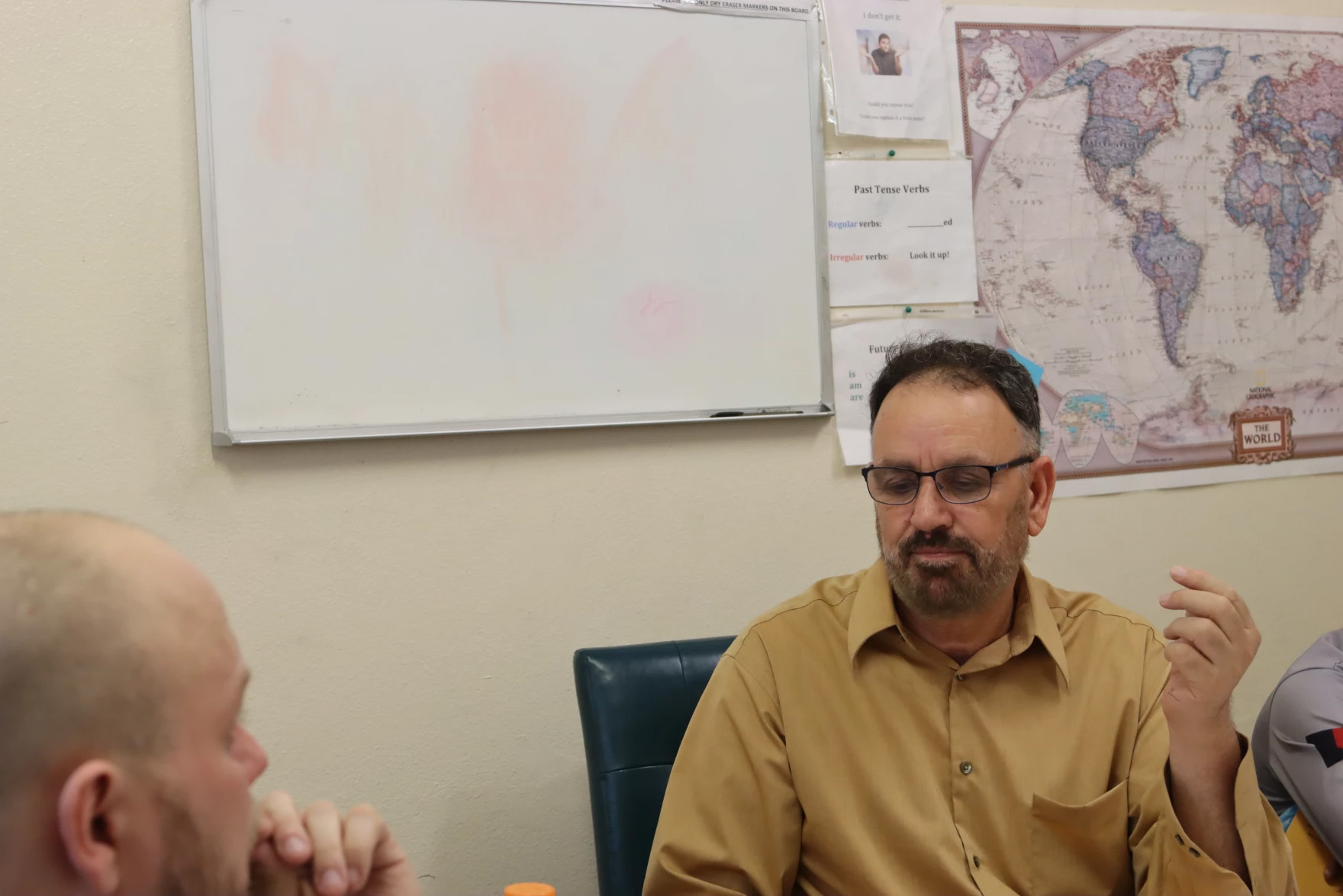 A man sits at a desk in conversation with another man across from him, seen in the foreground. Behind the man seated at the desk is a map of the world and whiteboard. This is Dr. Luqman Shah Sultani, and he is speaking with Alex Crous across from him.