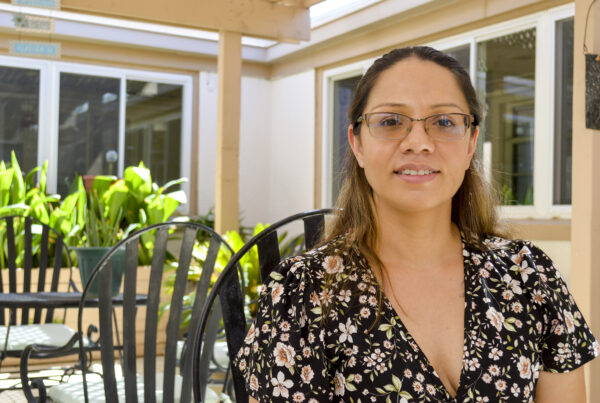 A woman sits in a a patio area outside on a sunny day. She is wearing a black floral dress and glasses and smiles at the camera.