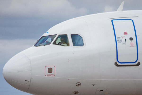 The cockpit of a commercial airplane is seen from the outside. I pilot wearing sunglasses and facemask looks out of the window.