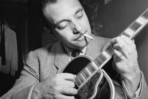 A black and white photo from the 1940s showing musician Django Reinhardt, a well-dressed man with slick-backed hair and thin mustache, playing a guitar while smoking a cigarette.