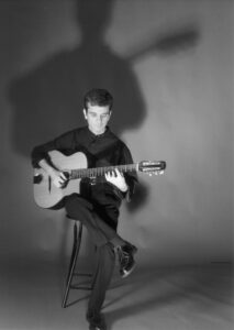 A black and white photo shows a man seated on a stool playing an acoustic guitar, the lighting casting a large shadow of himself behind him.