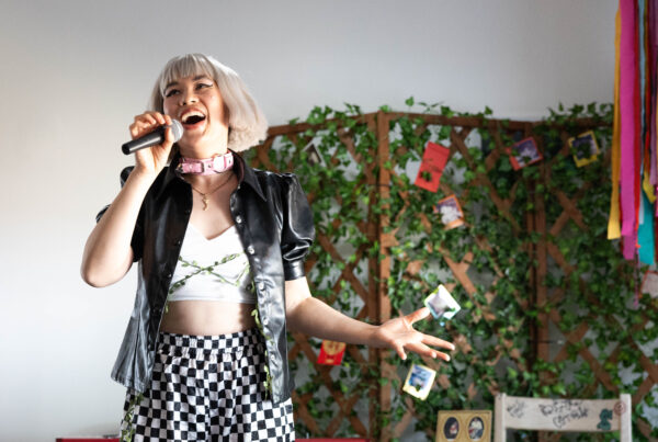 A smiling person sings into microphone. They are stylishly dressed in front of a background with greenery.