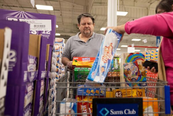 A man stands at a shopping cart full of boxes in a Sam's Club warehouse as a woman places another box into the cart