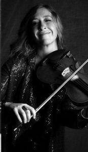 A black and white photo shows a woman smiling for the camera while holding her fiddle in a playing position.