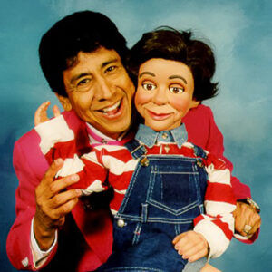 A man poses, holding up the arm of a puppet. This is Nacho Garcia with his puppet, Maclovio.