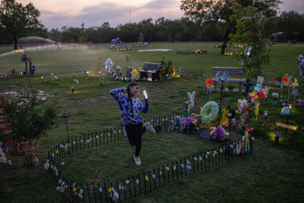 A young girl does a dance and sticks out her tongue while holding a smart phone. She is standing on the green grass in a graveyard.