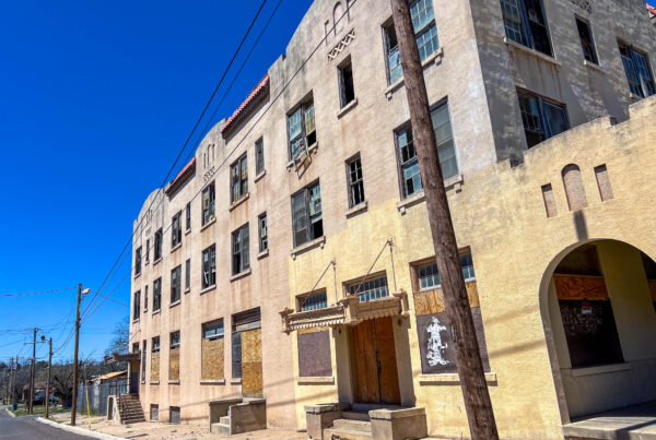 An old building is seen in the day time. It appears to be three stories, aged, and several windows are broken.