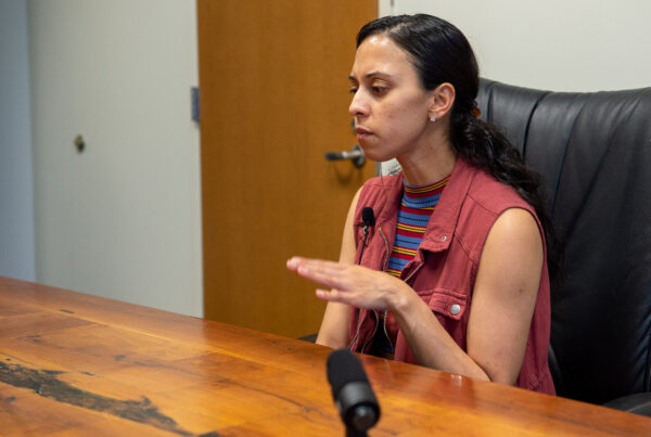 A photo of a woman at a wooden table in front of a microphone.