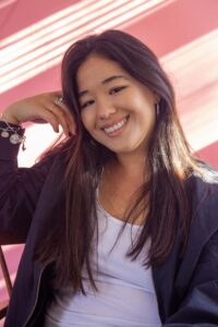 A photo of director Katherine Propper. She has long brown hair and is smiling at the camera. She is wearing a black casual jacket and a white shirt.