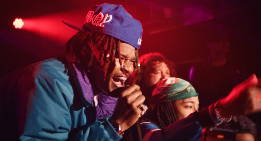 A photo of three or four people holding microphones close to their faces as they sing on a dark stage lit by red lights.