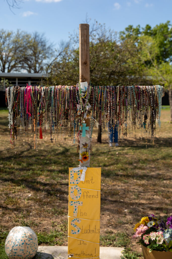 A photo of a small wooden cross with hundreds of rosaries hanging off of it. A sign underneath it reads "DOTS: Don't Offend The Students Do Something!"