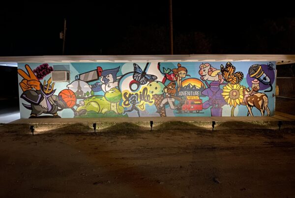 A small, rectangular building light from below at night. On the wall is a colorful mural with cartoon-like images of butterflies, mermaids, ice cream cones and other things many kids enjoy.