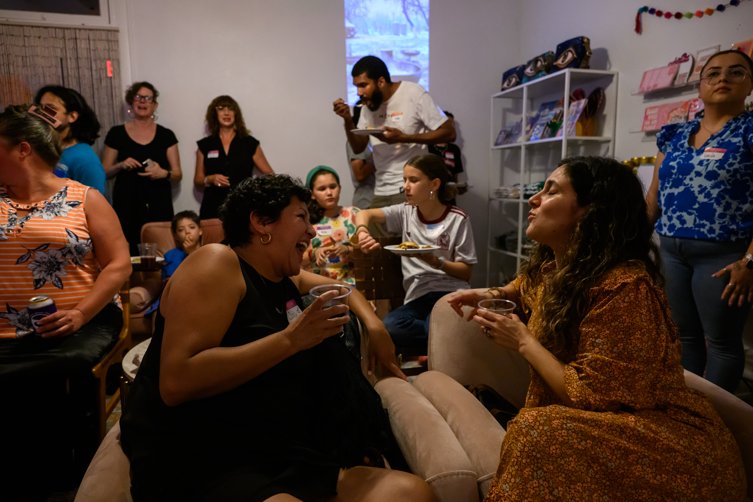Several people sit and stand as they eat and converse. Two people at the forefront of the image seem to be having an engaging conversation, as the woman to the right laughs raucously as her conversation partner speaks.