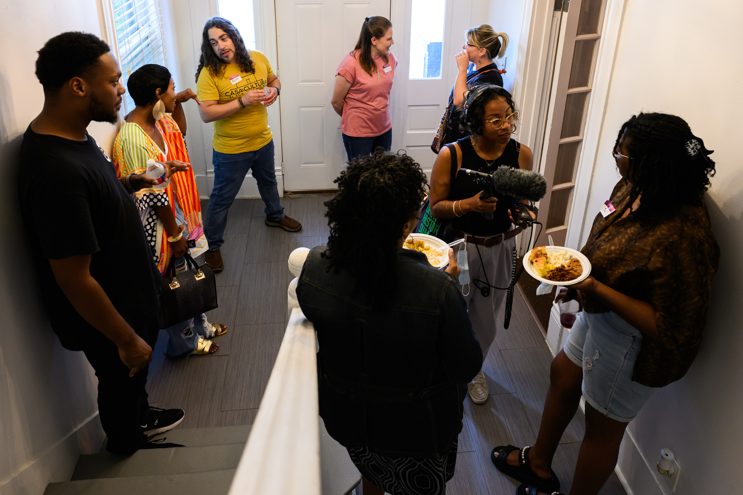 A group of people gather and converse in the entryway of a home, a stairway beside them. Zahra Crim, a KUT reporter, is scene holding a microphone and wearing headphones while interviewing two people holding plates of food.