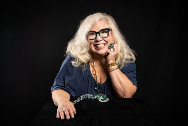 A blonde woman with big, black glasses smiles for the camera. She poses in front of a black background with a big necklace in front of her and a matching ring on her finger.