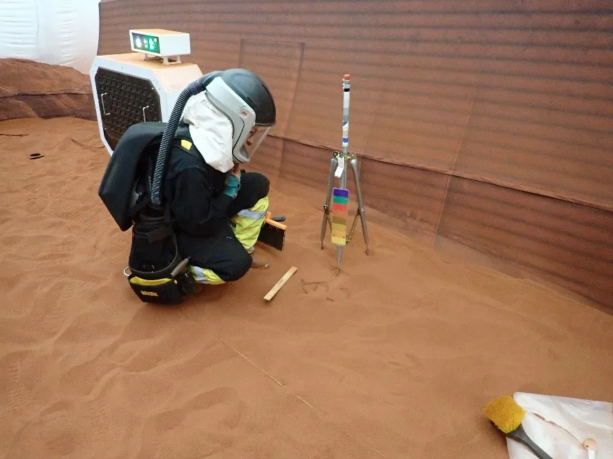 A person wearing a black and white space suit squats to examine equipment on a red dirt surface within a 3D printed enclosure.