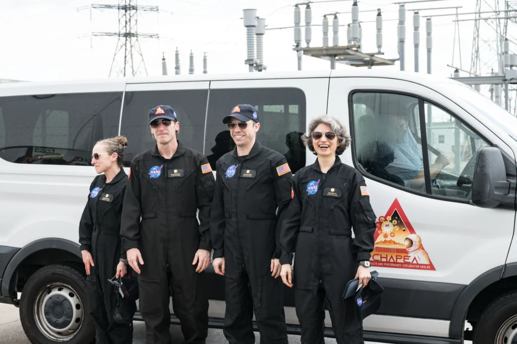 Four people stand outside of a white van. They are wearing black NASA work suits and sunglasses.