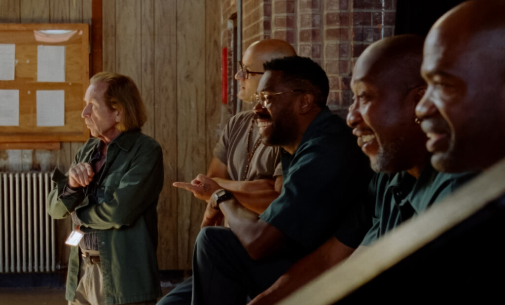 A still image from the film "Sing Sing" shows a row of people smile and observe a theatrical performance.