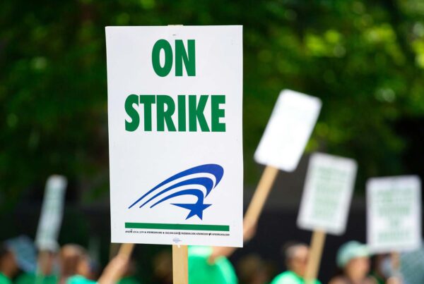 A white sign attached to a wooden stick that says "On Strike" in green letters