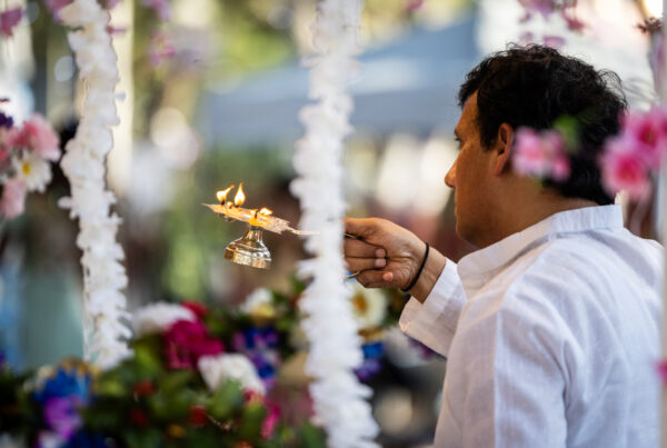 A man is seen from the side, holding a brass lamp with a small open flame on it. He's surrounded by strings of white, purple and pink flowers.