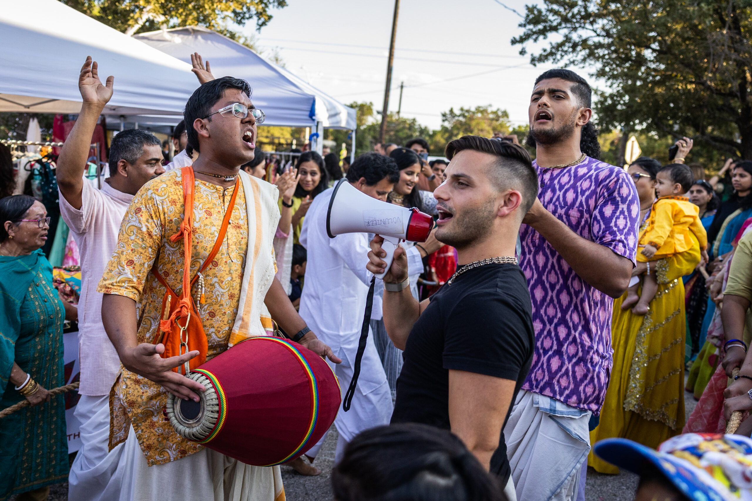 An animated crowd gathers two men leading a song, one singing into a bullhorn while another plays a hand drum.