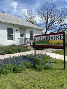 A vertical picture of the Calaboose museum sign and the small, white brick building behind it. The bluebonnets in the museum's front lawn are in bloom.