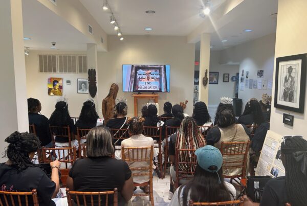A crowd of people sit in a gallery and face a screen with an image that reads "End Segregation."