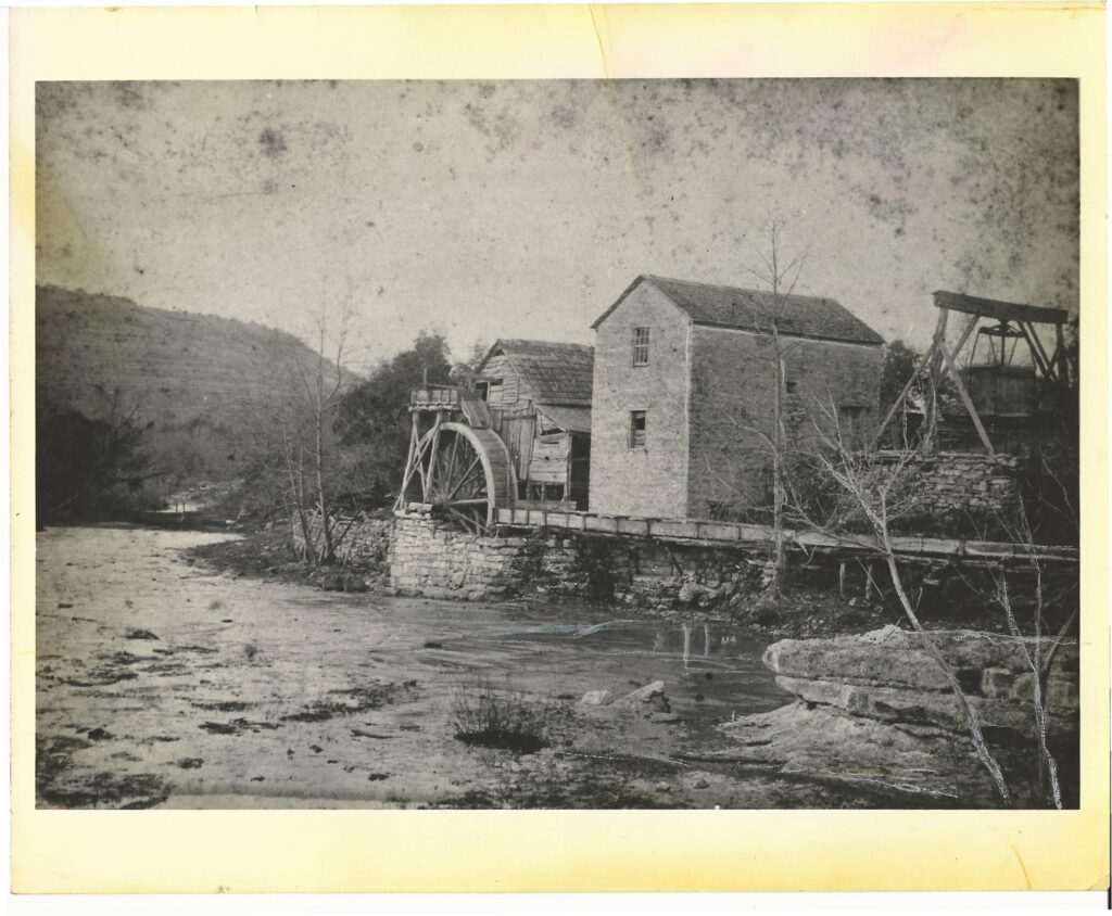 A black and white photos shows the original wood and stone mill with its wheel along a waterway.