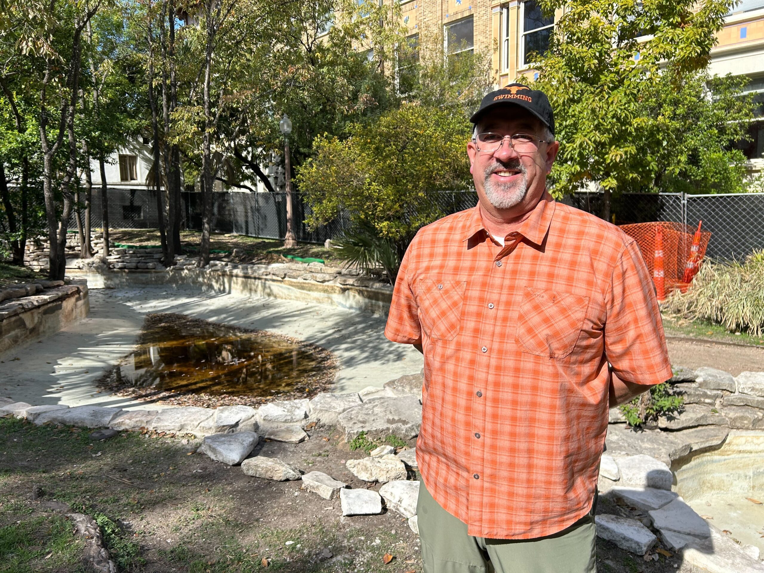 A person in an orange shirt smiles and stands in front of a nearly-empty shallow, pool-like pond.