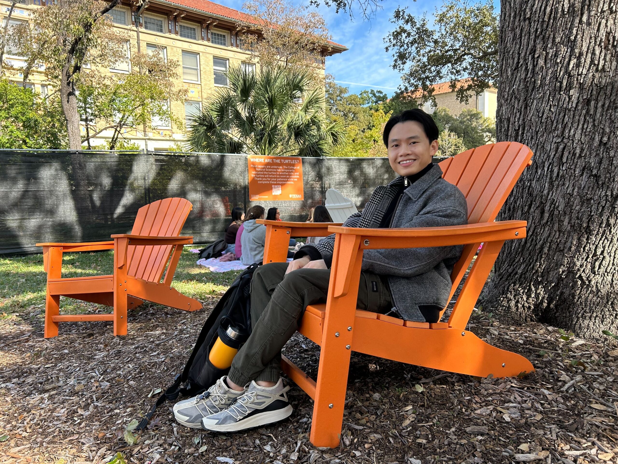 A person sitting in an orange Adirondack chair smiles for a photo. The chair is under a big oak tree. In the background is the temporary fence surrounding the UT Austin Turtle Ponds.