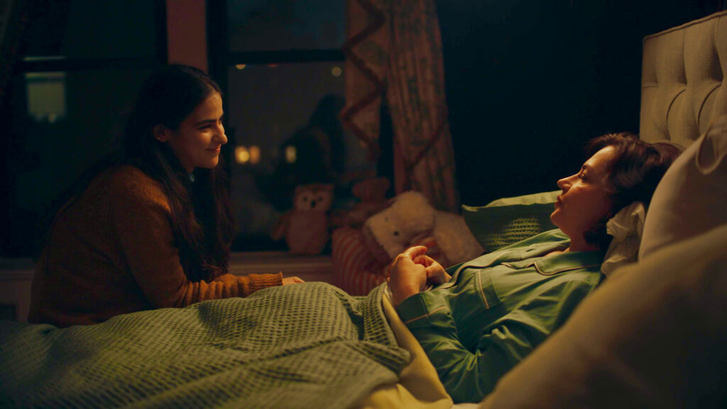 A film still shows a two smiling women -- one sitting next to bed and the other lying in the bed.