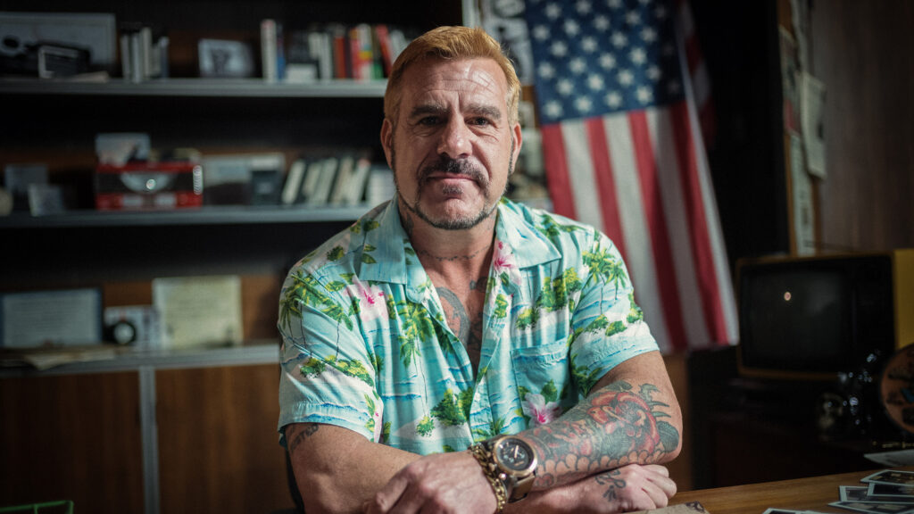 A film still shows a man wearing a Hawaiian shirt and sitting at a desk in front of an American flag.