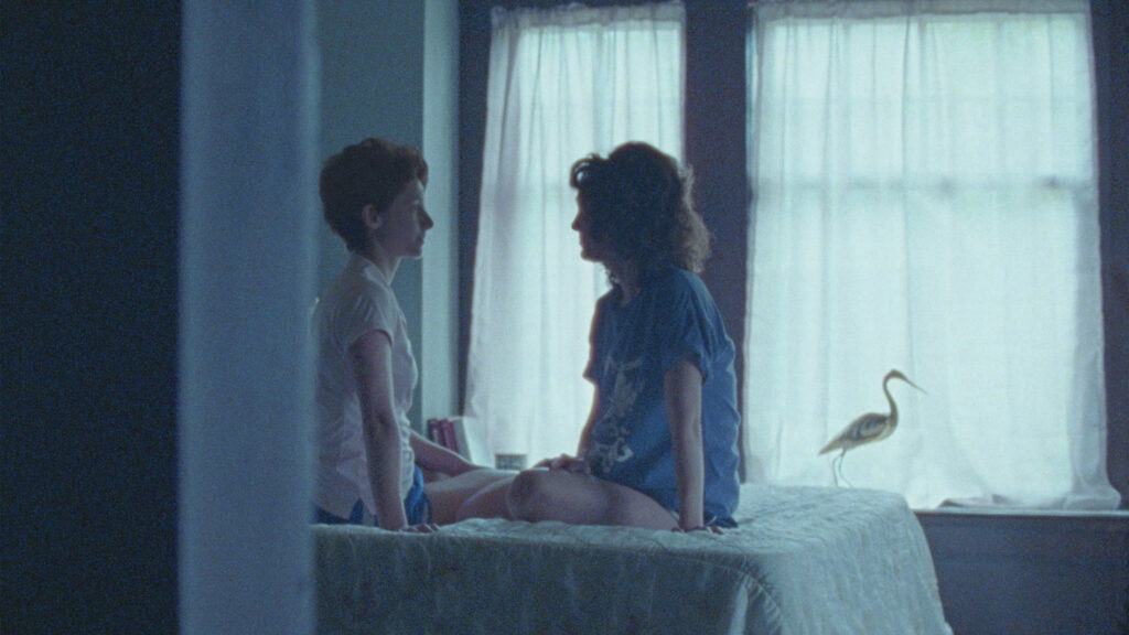A film still shows two women wearing t-shirts and looking at one another while sitting on a bed.