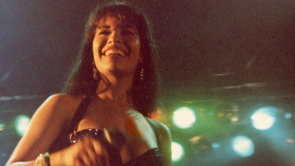 A film still shows Selena Quintanilla smiling on stage during a performance.