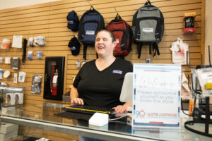 A woman in a black v-neck shirt stands at a register behind a counter, in front of a wall of backpacks.