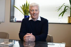 A portrait of a man wearing a dark blue sweater over a white collared shirt. He's sitting down with his hands resting on the table in front of him.