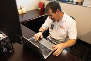 A man in a white polo shirt sits at a desk working on a laptop.