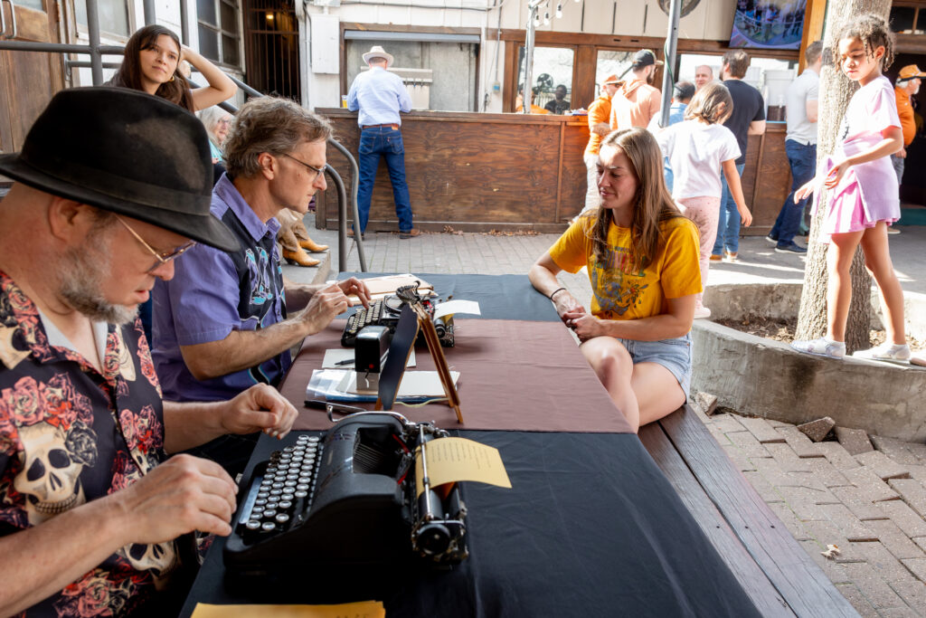 Two people type poems on vintage typewriters at an outdoor picnic table.