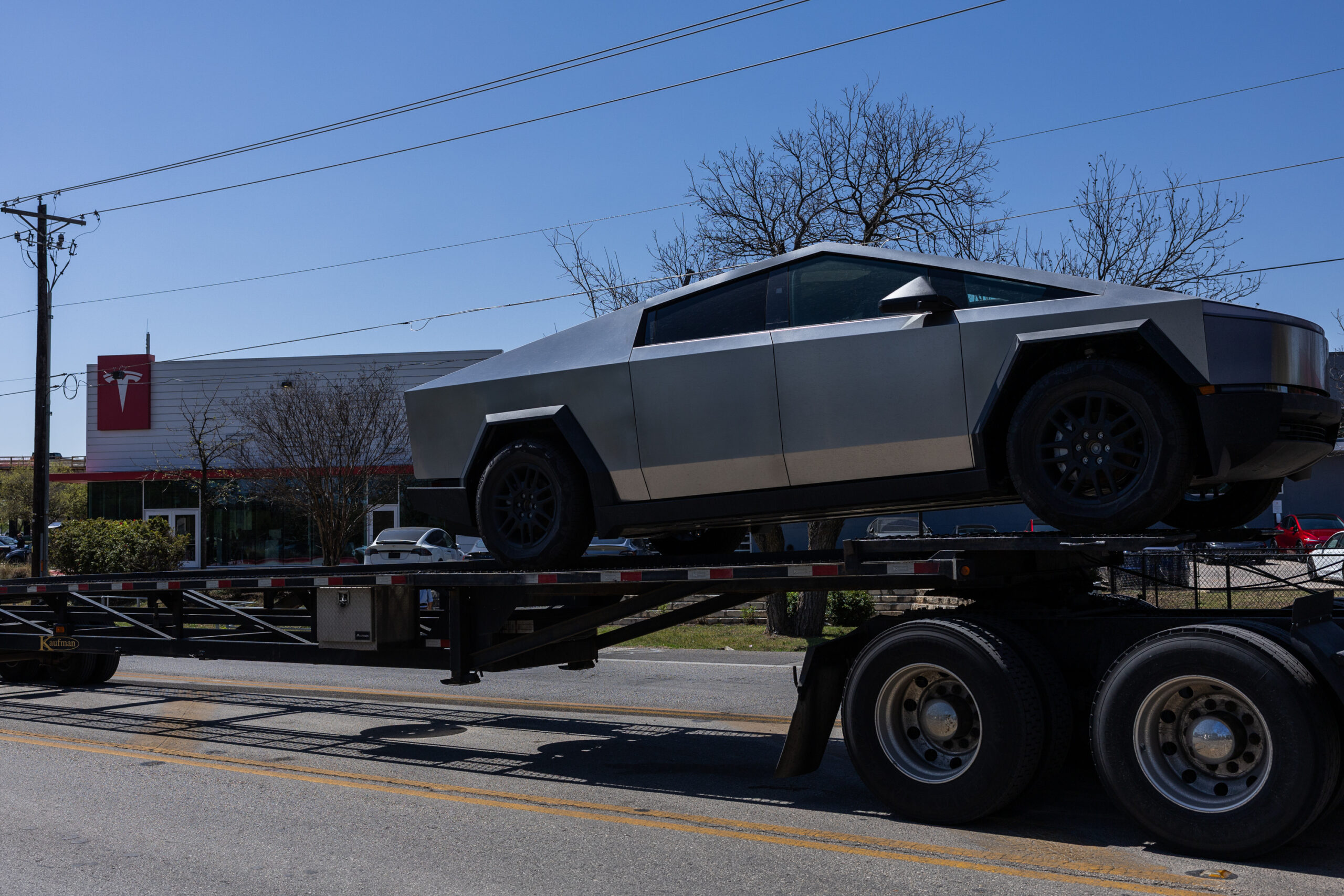 A Tesla cybertruck is seen in the back of a large flatbed truck in front of a Tesla dealership.
