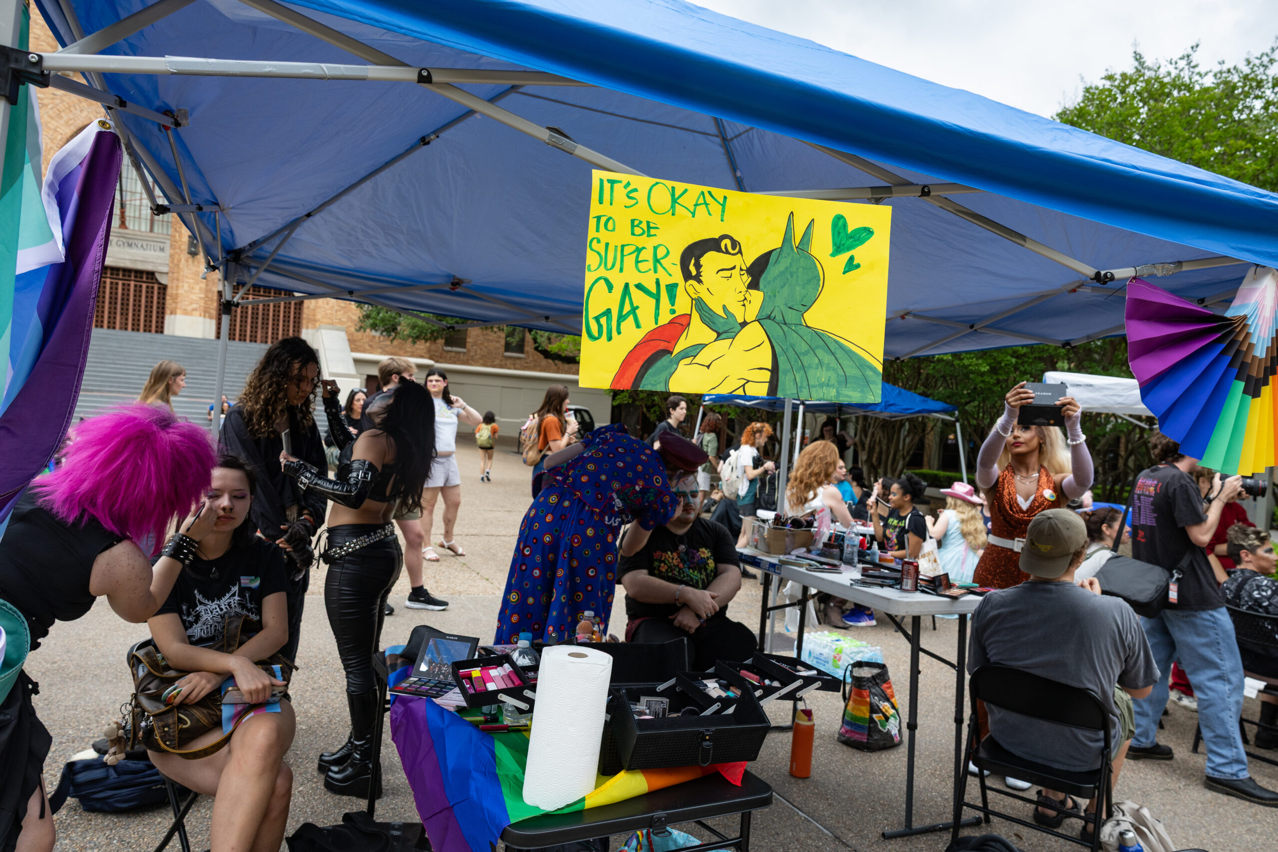 Several students are seated under a popup canopy outdoors as they have their makeup done by drag queens. A sign hanging from the canopy depicts comic book heroes Superman and Batman kissing with the phrase 