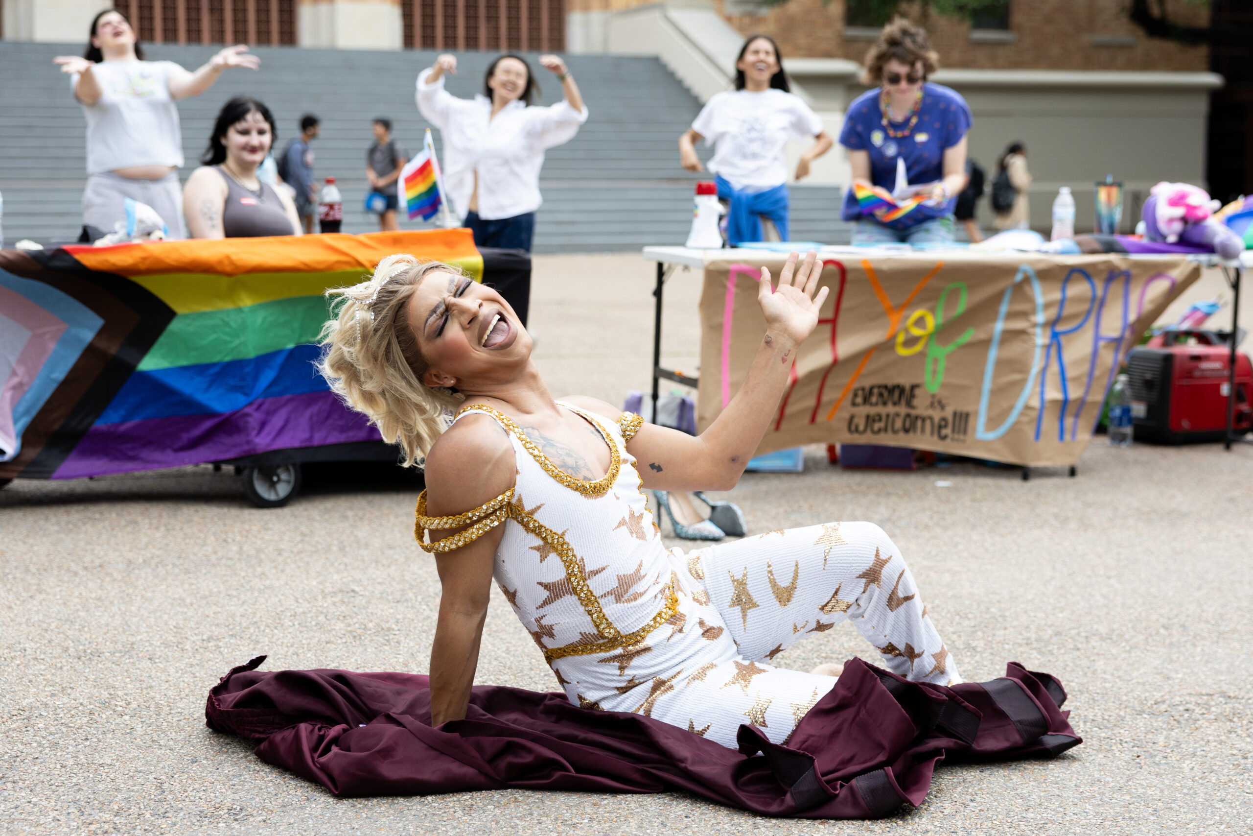 A drag queen strikes a pose on the ground as they lip sync with their eyes closed. Behind them, students cheer on at tables with signs reading 