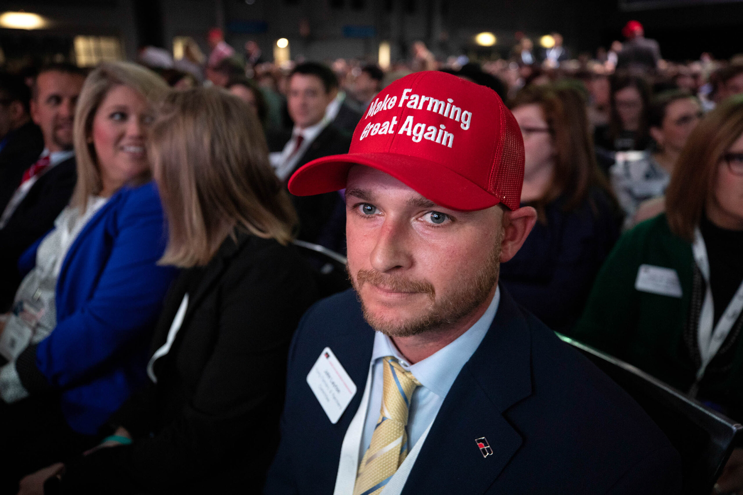 A young man is seen sitting in a audience. He is dressed in a suit, wearing a red baseball hat reading 