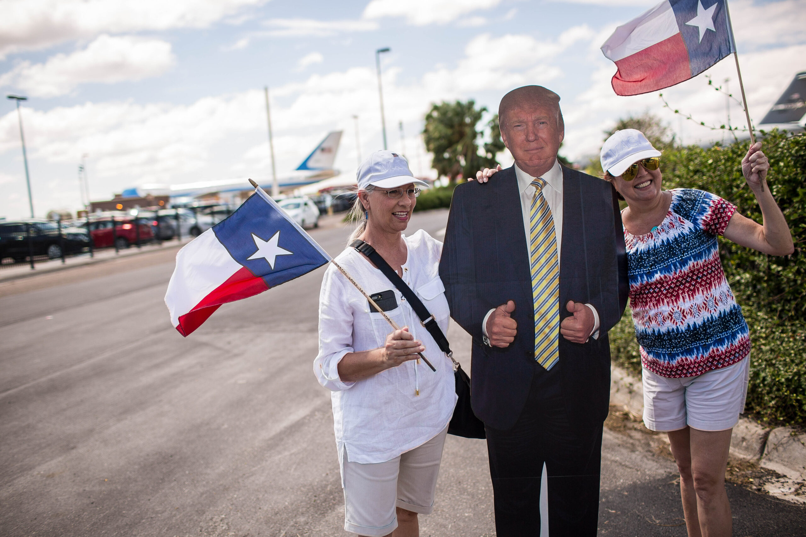 Two women holding Texas flags pose outdoors with a life-sized cardboard cutout of President Donald Trump. Air Force One is seen on the tarmac in the background.
