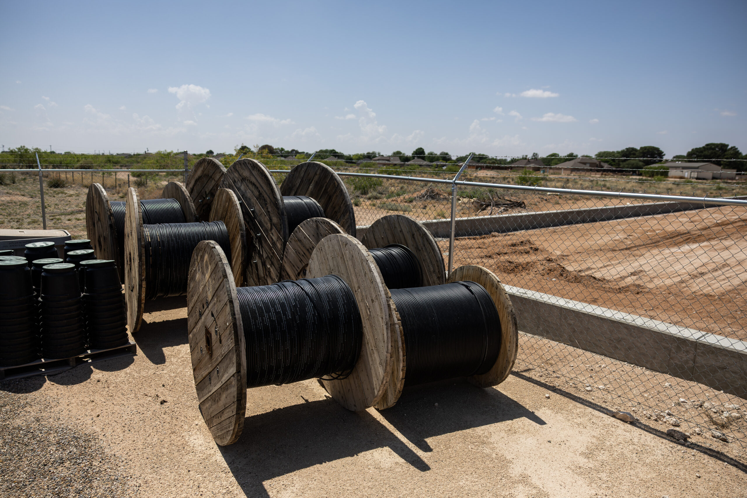 Large spools of black wires are seen resting against a wire fence outdoors.