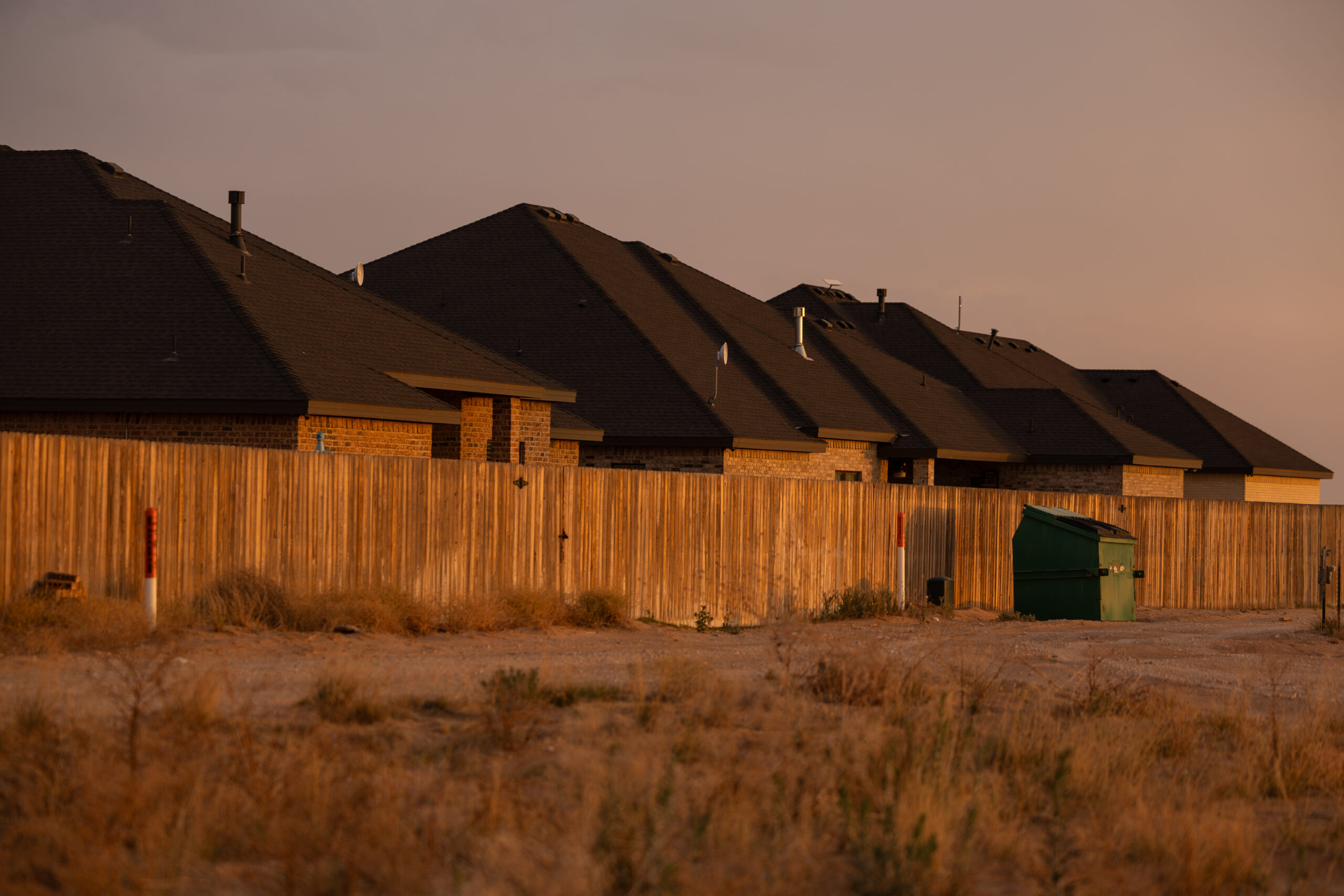 A row of homes are seen behind wooden privacy fences. A couple of orange and white poles designating where fiber optic cables are buried stand along a dirt walkways along the fence.