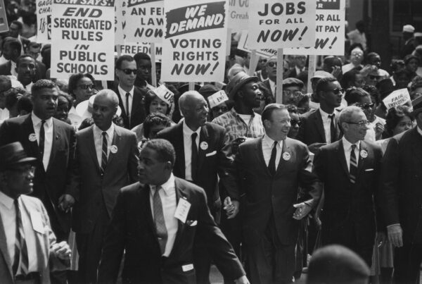 A black and white photo of a crowd of men in suits carrying signs such as "we demand voting rights now!"