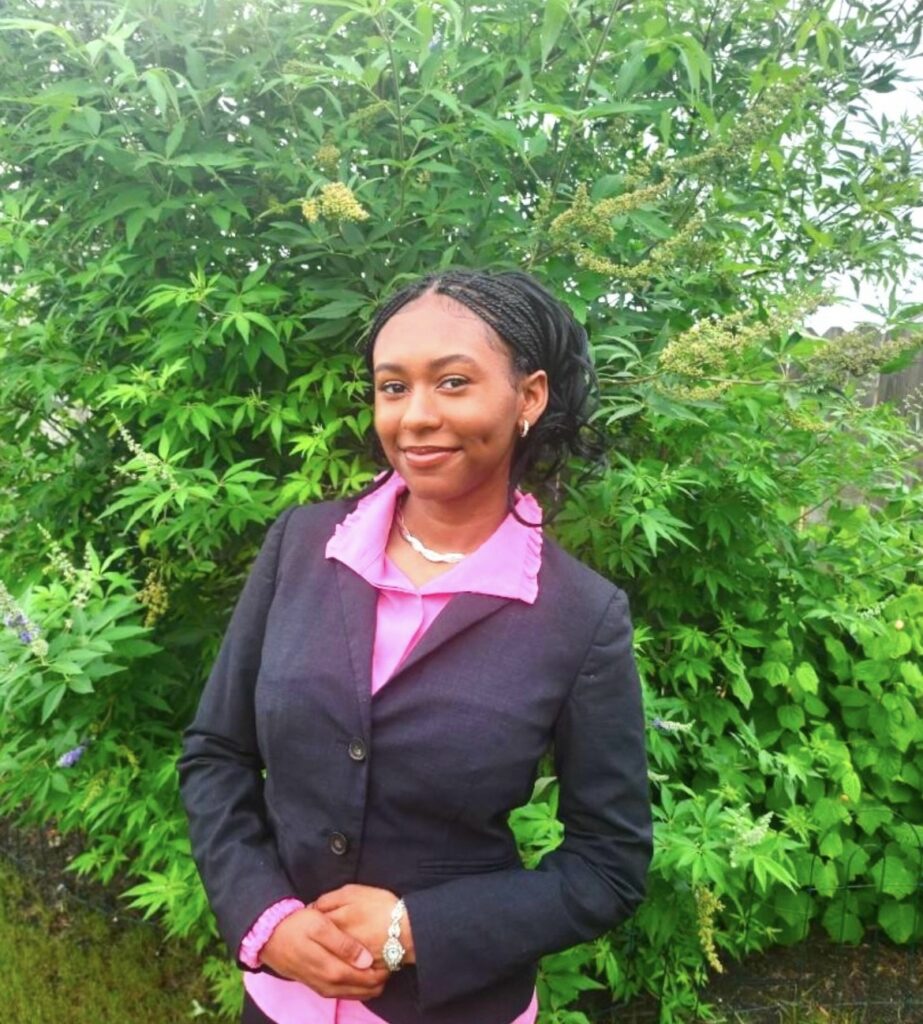 A young woman wearing a suit jacket poses in front of greenery.