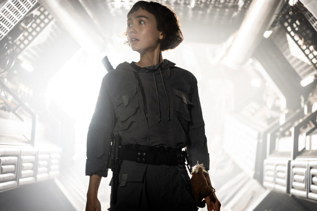 A still image from the series shows a young woman with short brown hair standing in front of a bright white futuristic background.